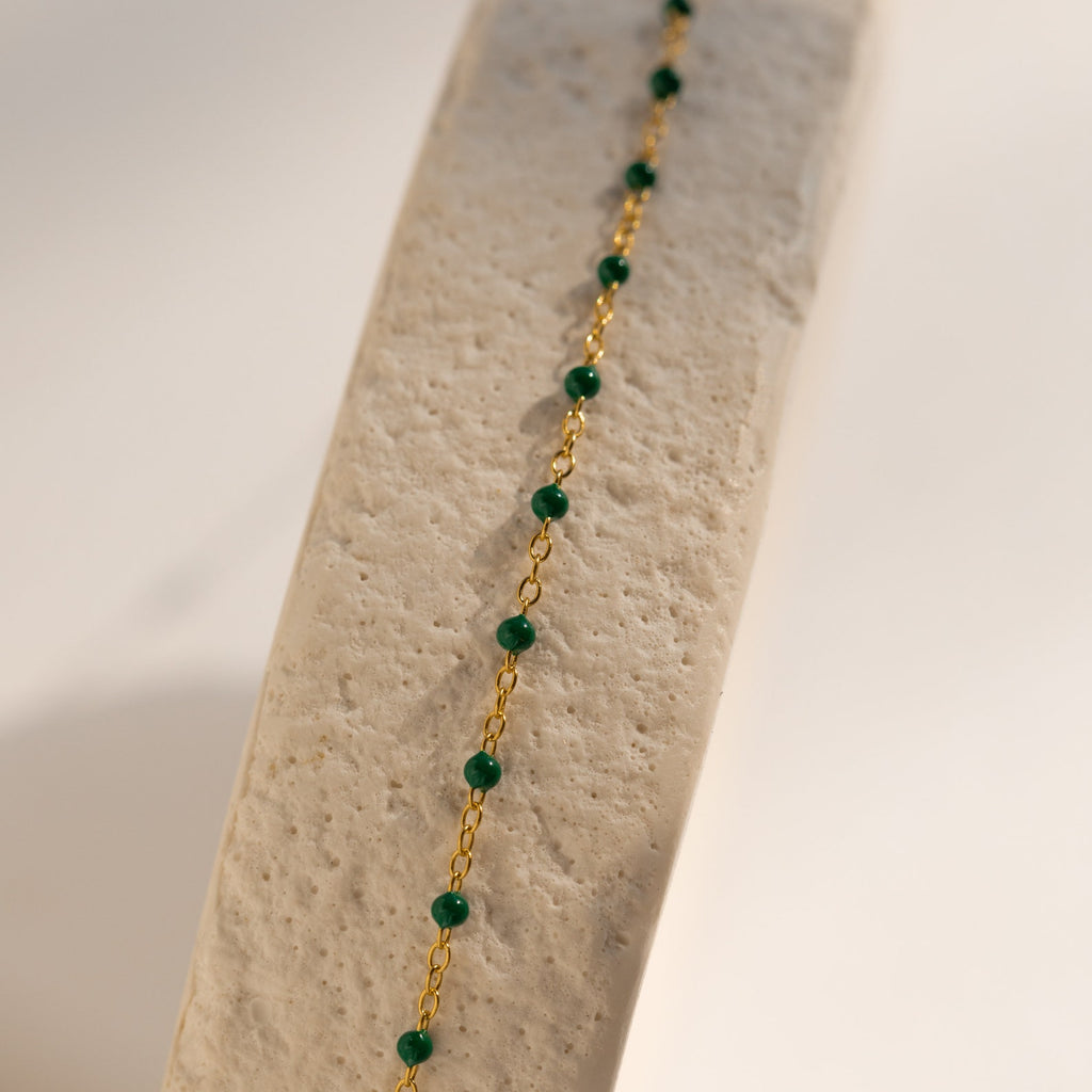 The Emerald Station Necklace features gold chain and emerald green beads, elegantly draped over a textured beige surface in soft light—a delicate beaded boho accessory for subtle elegance.