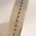 The Emerald Station Necklace features gold chain and emerald green beads, elegantly draped over a textured beige surface in soft light—a delicate beaded boho accessory for subtle elegance.