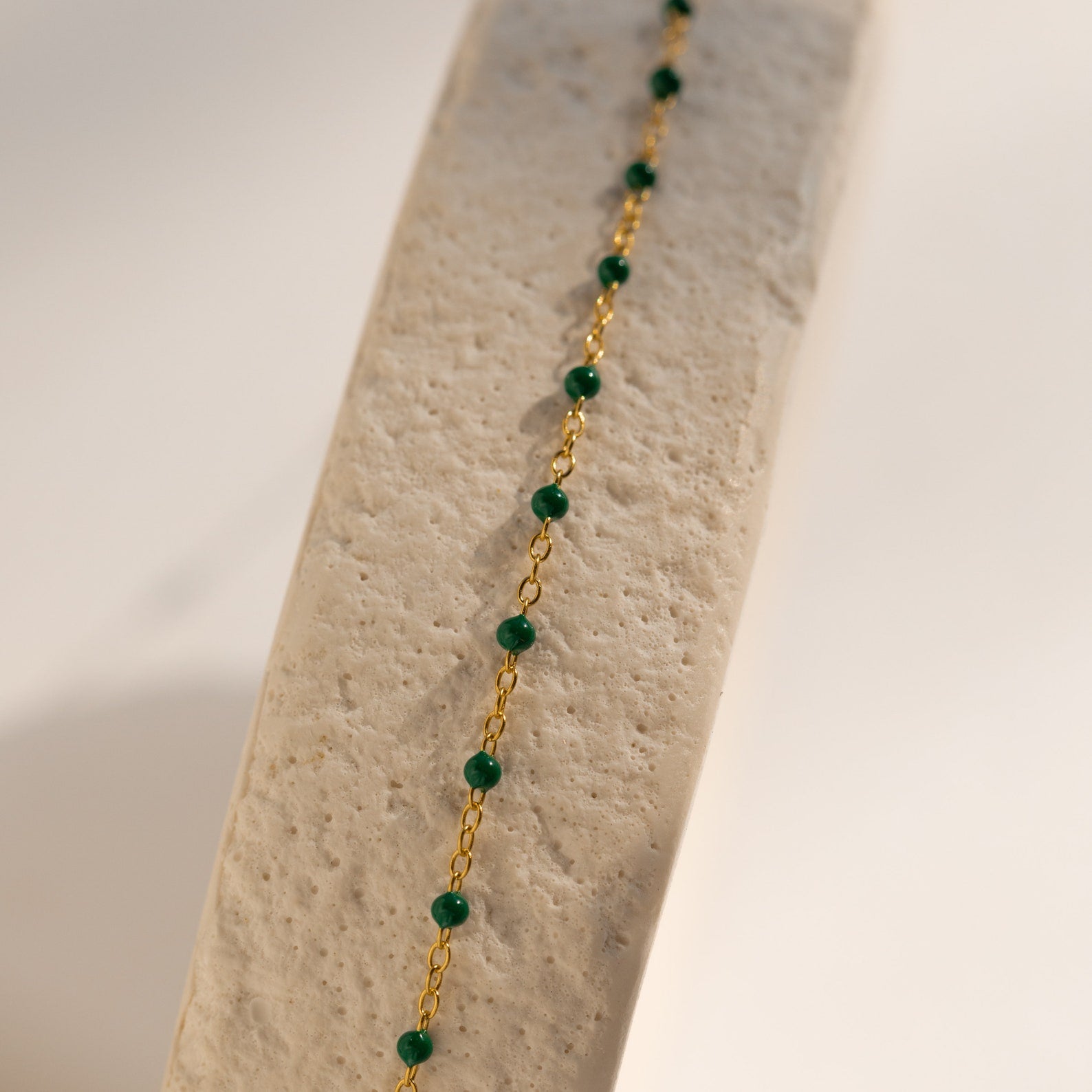 The Emerald Station Necklace features gold chain and emerald green beads, elegantly draped over a textured beige surface in soft light—a delicate beaded boho accessory for subtle elegance.