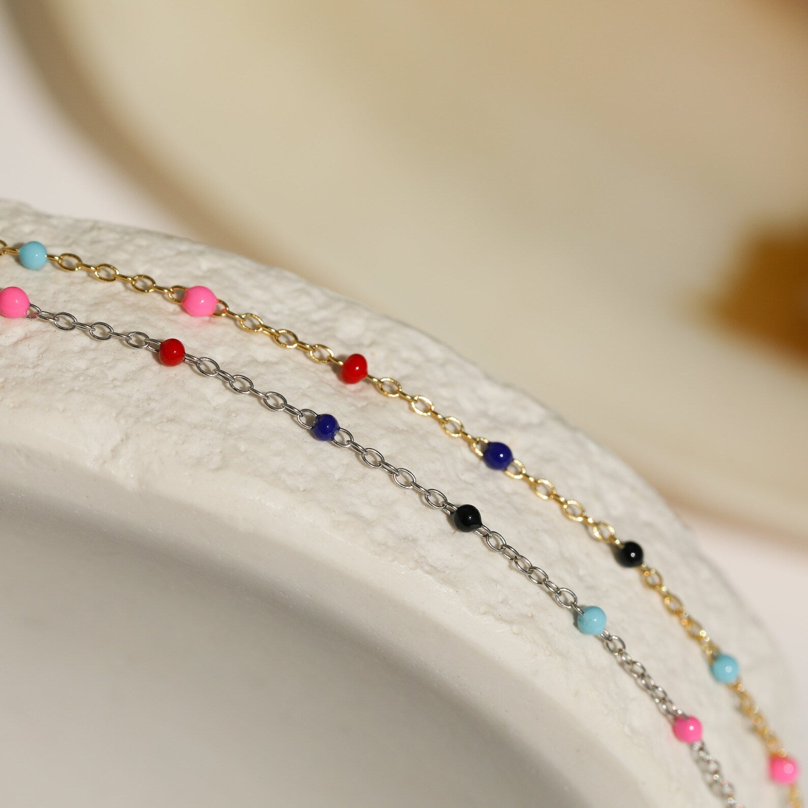 The Rainbow Station Necklace, with its delicate chain and colorful beads, rests on the rim of a textured white ceramic bowl, evoking the charm of a vibrant beaded accessory.