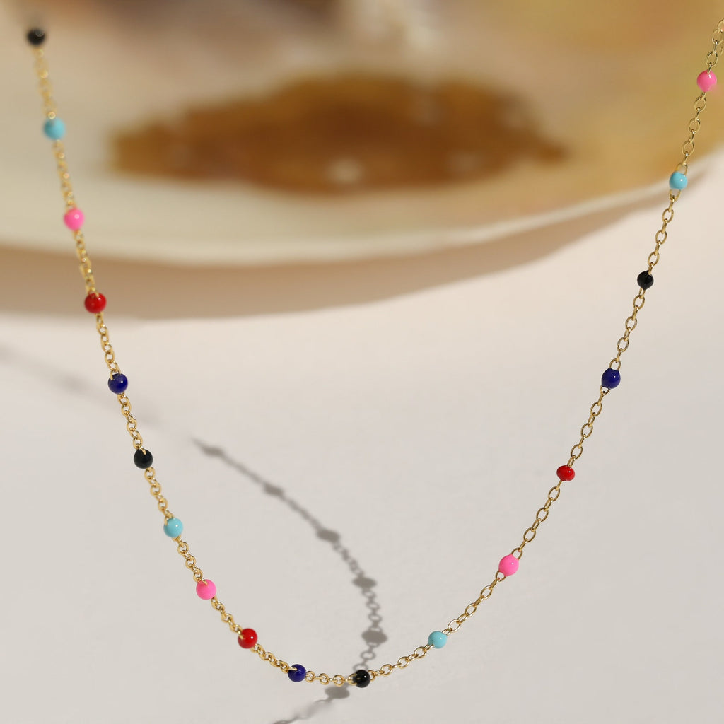 The Rainbow Station Necklace features a gold chain adorned with small colorful beads in pink, blue, red, and black—an ideal playful accessory for anyone looking to add a touch of rainbow charm.