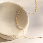The White Station Necklace, a delicate gold chain with dainty white beads, is elegantly displayed on a textured white ceramic dish under soft, natural light.