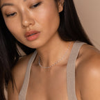 Woman with long dark hair wears a beige knit top and the White Station Necklace, a dainty piece featuring white beads, as she looks down.