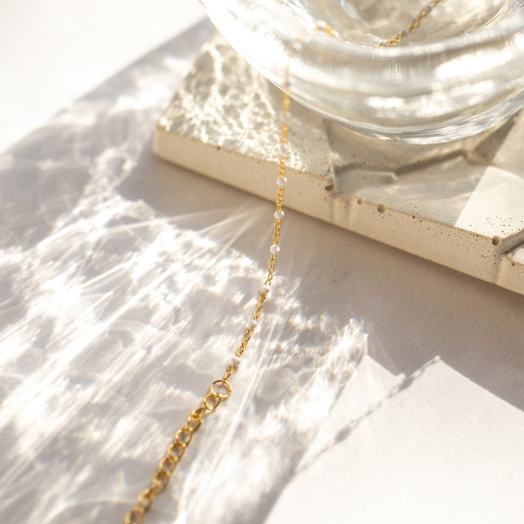 The White Station Necklace features a delicate gold chain with dainty white beads, displayed on a textured stone tile as light plays nearby—an elegant handmade piece perfect for any occasion.