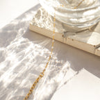The White Station Necklace features a delicate gold chain with dainty white beads, displayed on a textured stone tile as light plays nearby—an elegant handmade piece perfect for any occasion.