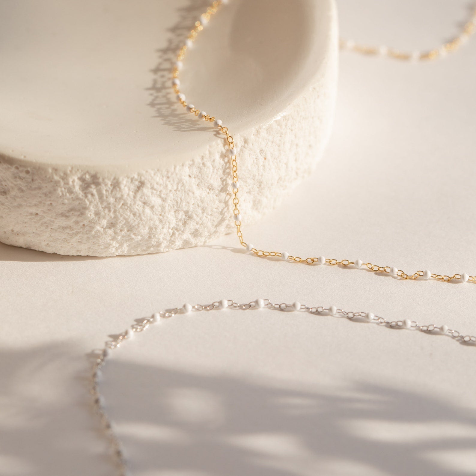 The White Station Necklace features delicate gold and silver chains with dainty white beads, displayed on textured white stone against a soft, neutral background.