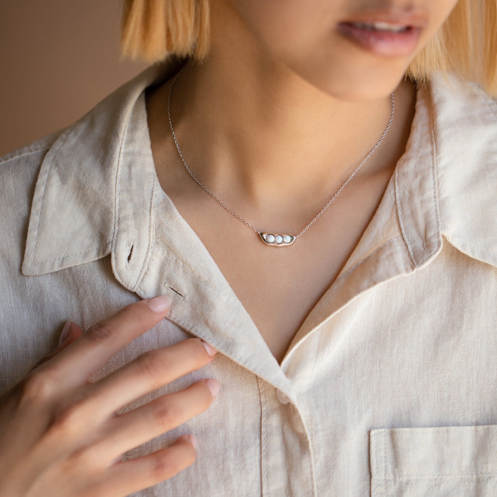 A woman in a cream shirt gently touches her collar, showcasing the Peas in a Pod Necklace with its three-pea pod pendant for a meaningful, stylish accent.