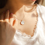 A woman wearing a white top and a silver Pearl Disc Locket Necklace with a round, pearly pendant resting on her collarbone—a meaningful gift that can hold special memories like a photo.