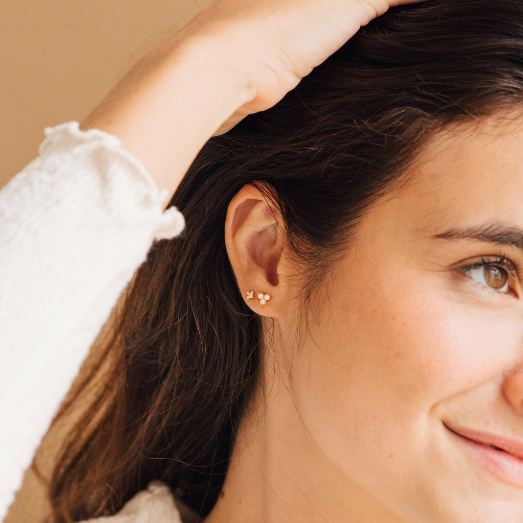 A woman with brown hair wears the Maud Pearl Studs—small gold flower-shaped earrings—smiling and touching her head for an effortlessly elegant, timeless gift look.