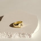The Traditional Initial Heart Signet Ring, engraved with the letter R, is shown on a textured white stone surface—a timeless personalized ring and an ideal anniversary gift.