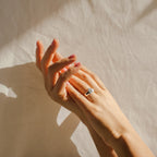 Two hands with manicured nails, one adorned with the Traditional Initial Heart Signet Ring, rest against a softly lit beige fabric background.