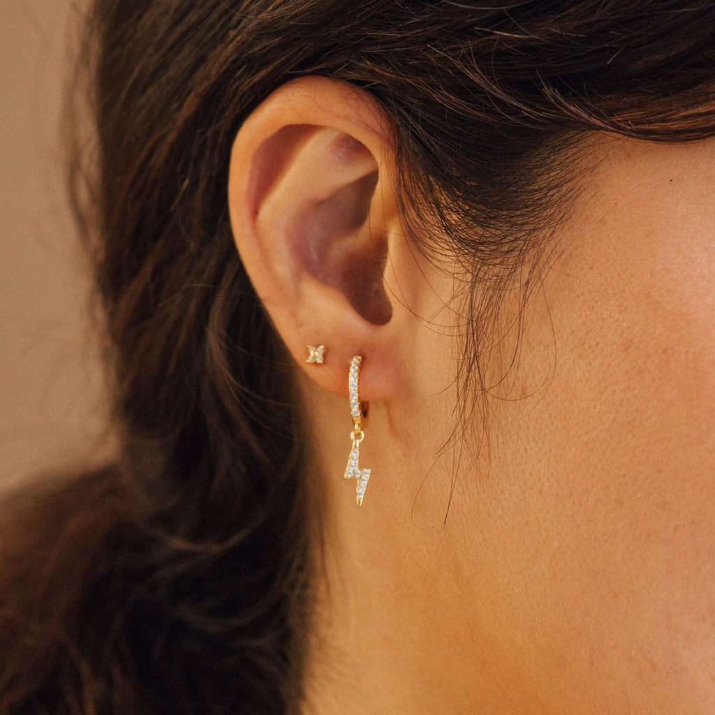 Close-up of an ear with two gold earrings: a small stud and the stylish Striker Pave Huggies—a dangling lightning bolt set with sparkling crystals.