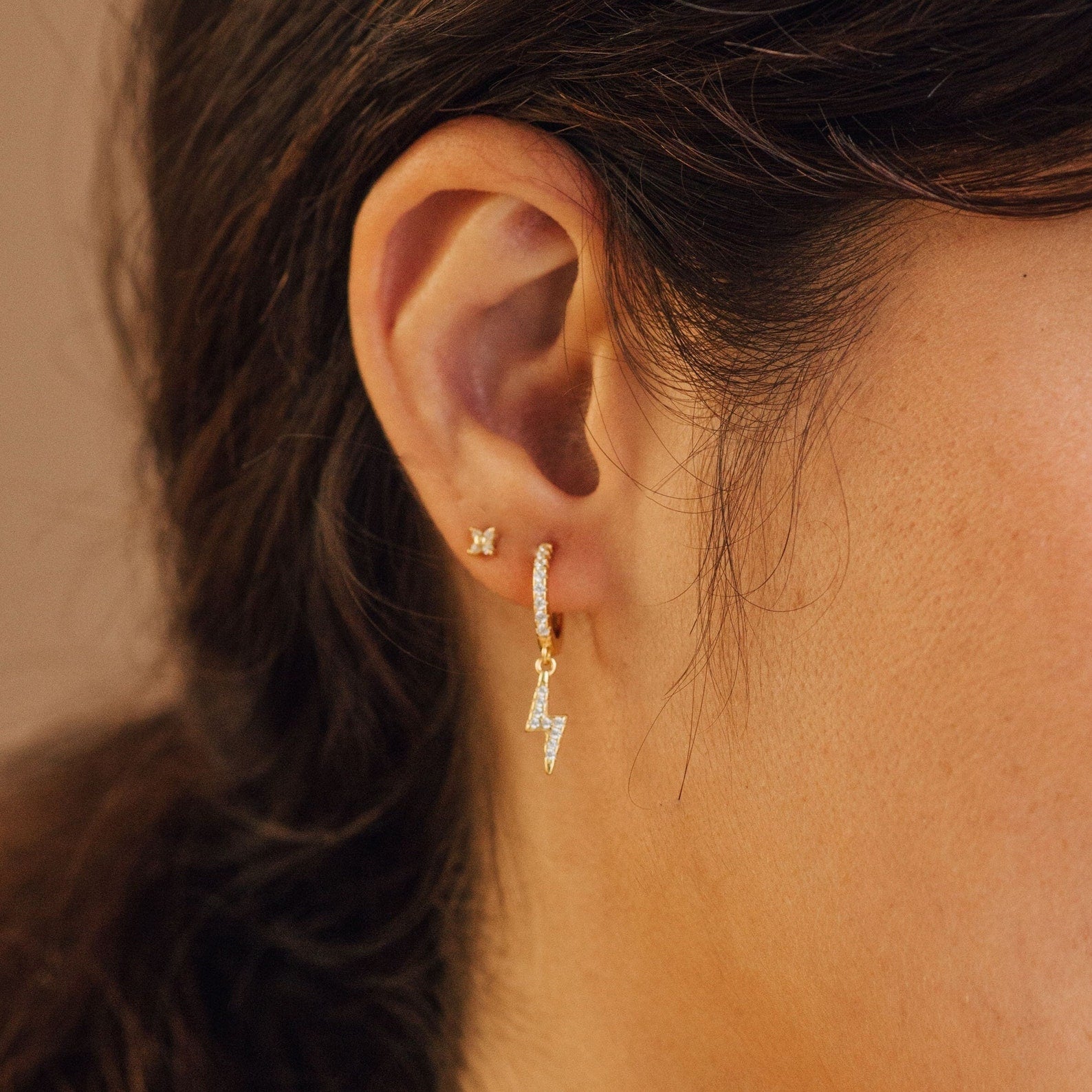 Close-up of an ear with two gold earrings: a small stud and the stylish Striker Pave Huggies—a dangling lightning bolt set with sparkling crystals.