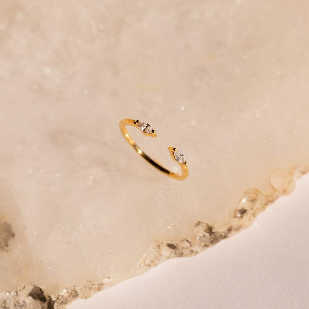 The Celeste Duo Marquise Ring features a gold open design with two small clear gems, beautifully presented on a light beige textured surface.