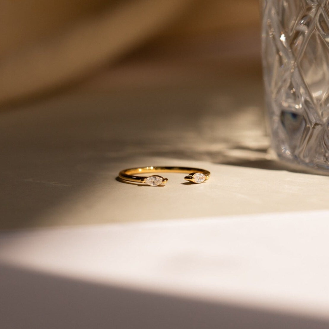 Celeste Duo Marquise Ring with two teardrop-shaped gemstones beside textured glass on a sunlit surface, highlighting its elegant open ring design.