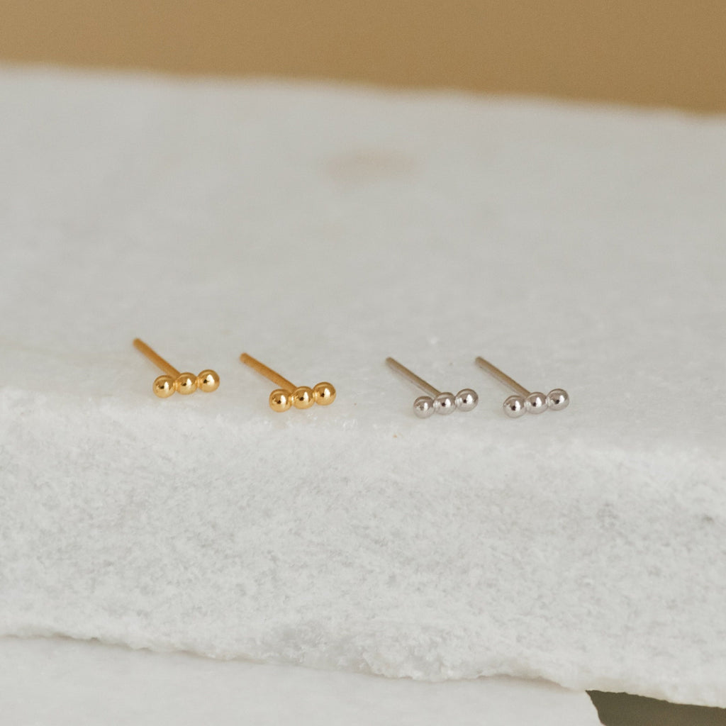Two pairs of Trio Beaded Bar Studs—one gold, one silver—with three small round beads each, rest on a white surface. These minimalist earrings are perfect for everyday wear.