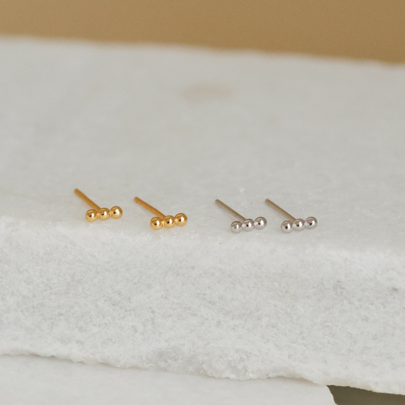 Two pairs of Trio Beaded Bar Studs—one gold, one silver—with three small round beads each, rest on a white surface. These minimalist earrings are perfect for everyday wear.