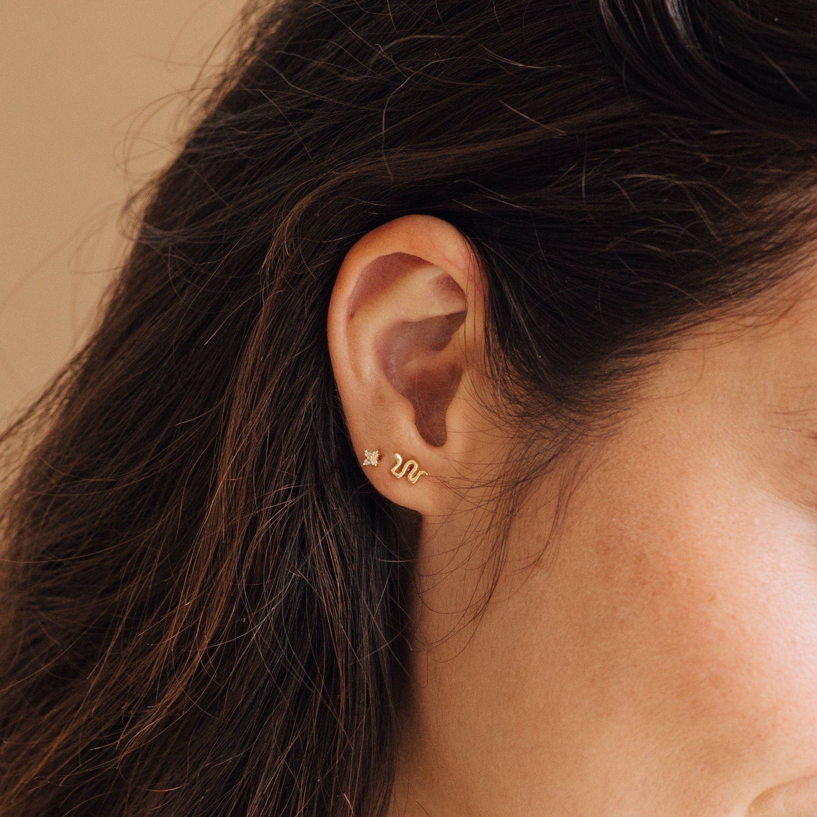 A close-up of a woman's ear wearing dainty jewelry: the gold Tiny Python Studs—a small, snake-shaped earring—paired with a star stud, perfect for adding boho-luxe style to your earrings collection.