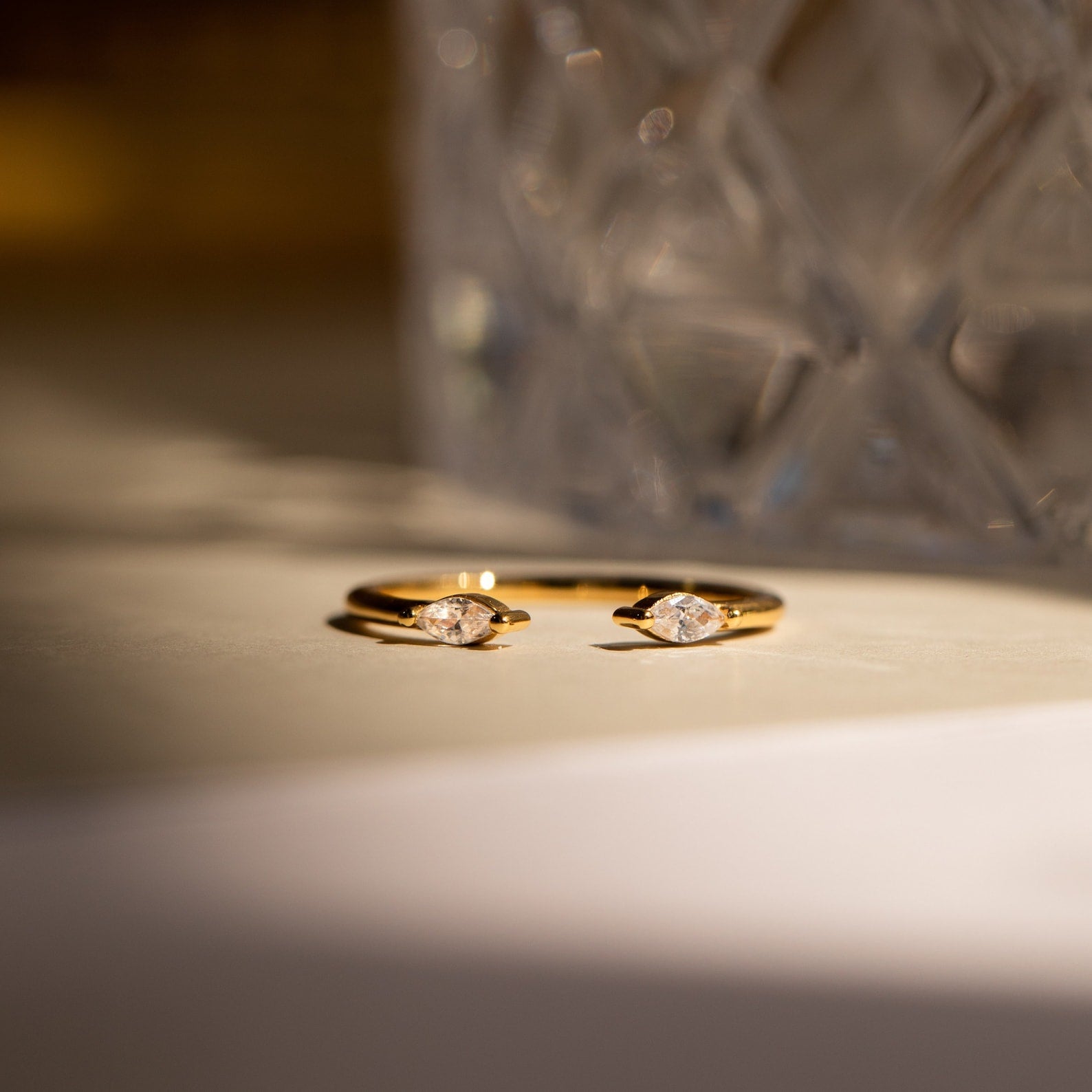 The Celeste Duo Marquise Ring showcases two marquise-cut diamonds on a gold open band, displayed on a light surface with a blurred glass object in the background.