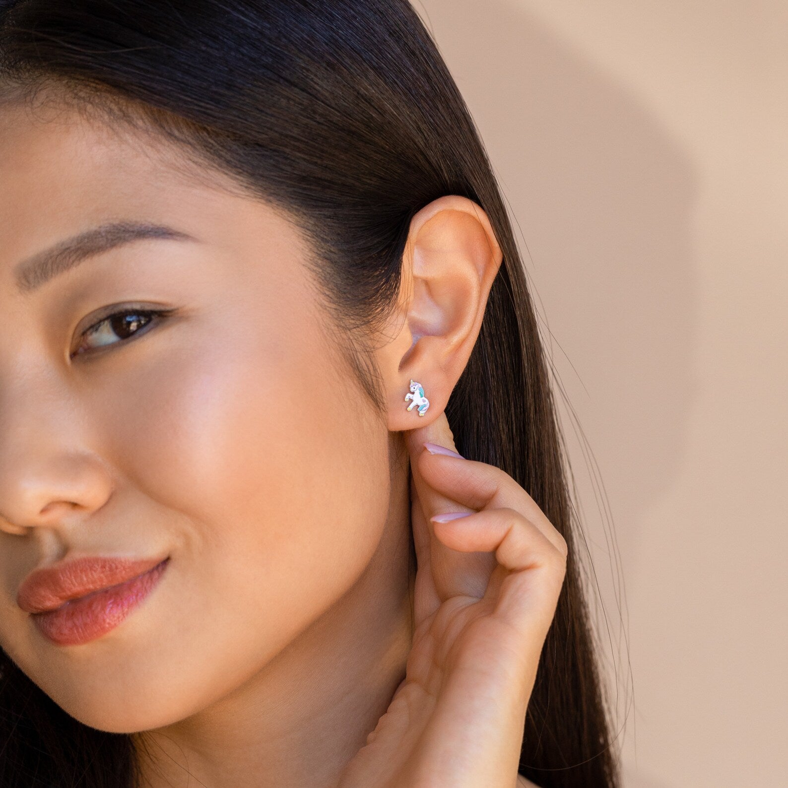 A woman touches her ear, showcasing Unicorn Studs—charming pastel enamel earrings.