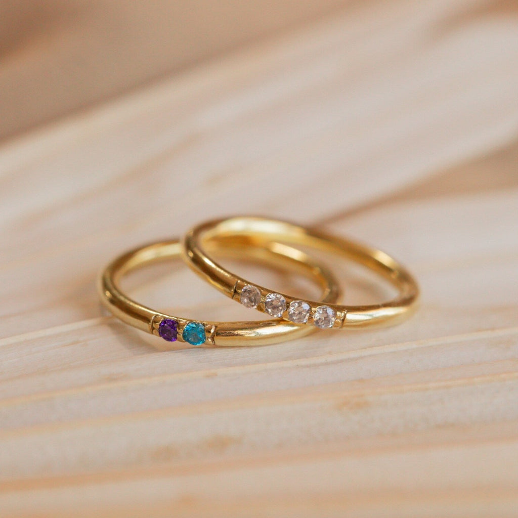 A Mini Birthstone Ring in Rose Gold, featuring three colored gemstones, and another gold ring with four clear stones rest elegantly on a wooden surface.