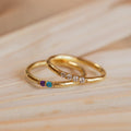 A Mini Birthstone Ring in Rose Gold, featuring three colored gemstones, and another gold ring with four clear stones rest elegantly on a wooden surface.