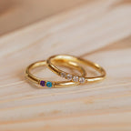 A Mini Birthstone Ring in Rose Gold, featuring three colored gemstones, and another gold ring with four clear stones rest elegantly on a wooden surface.
