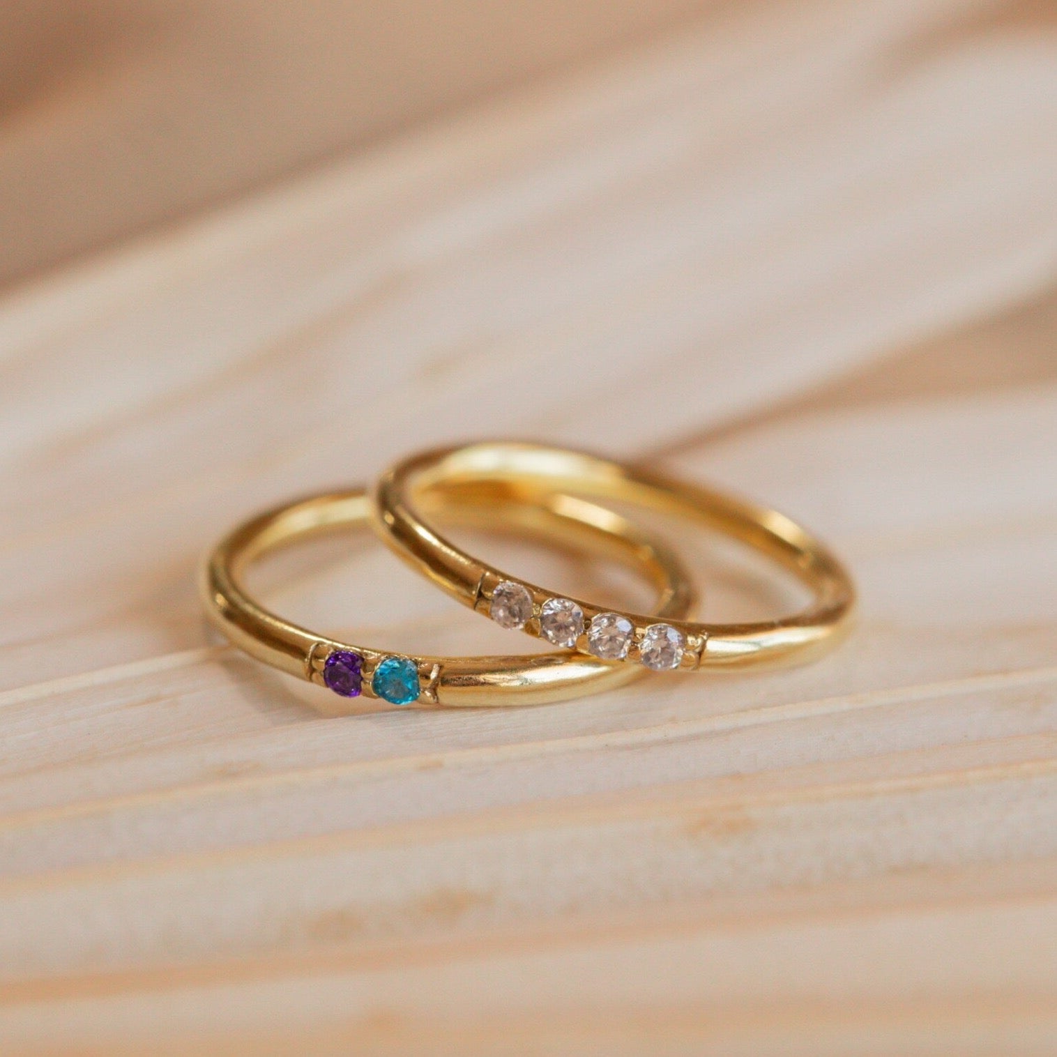 A Mini Birthstone Ring in Rose Gold, featuring three colored gemstones, and another gold ring with four clear stones rest elegantly on a wooden surface.