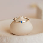 The Mini Birthstone Ring in Sterling Silver, featuring a petite blue stone, is elegantly displayed on a smooth, round ceramic stand.