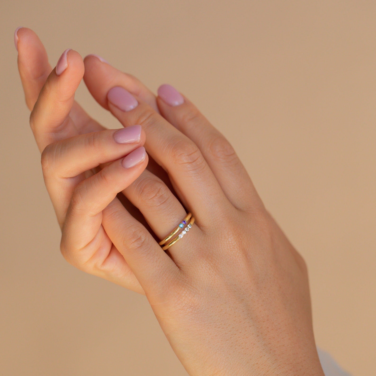 A hand with light pink nails wears the Mini Birthstone Ring and a gold band with small diamonds, set against a beige background.