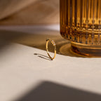 The Serafina Diamond Ring, a delicate gold band with two sparkling diamonds, sits beside a ribbed amber glass on a sunlit surface.