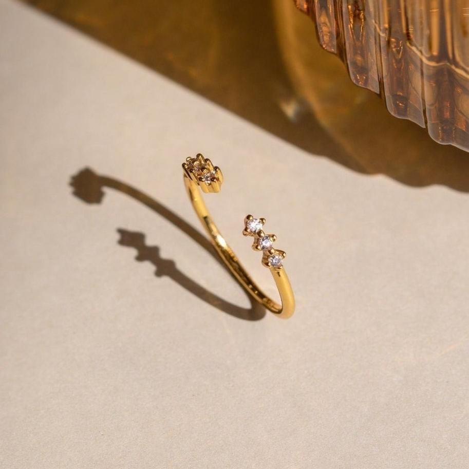 The Serafina Diamond Ring, a dainty gold open ring adorned with three small diamonds, casts a shadow on a light surface beside a textured glass object.