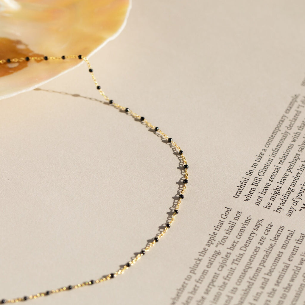 The Black Station Necklace, featuring delicate gold and black charms, rests on a shell atop a printed page—a stylish nod to today's fashion trends.