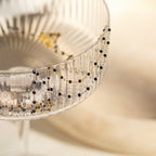 The Black Station Necklace, featuring delicate gold and black beads, is draped over the rim of a textured glass bowl.