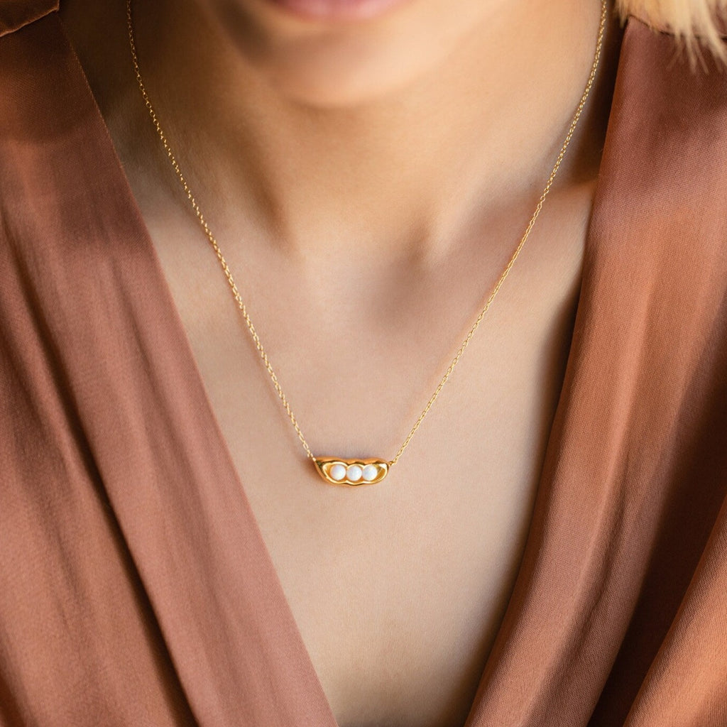 A woman wears the Peas in a Pod Necklace—a gold pendant with three pearls—paired with a brown V-neck top, highlighting meaningful jewelry.