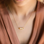 A woman wears the Peas in a Pod Necklace—a gold pendant with three pearls—paired with a brown V-neck top, highlighting meaningful jewelry.