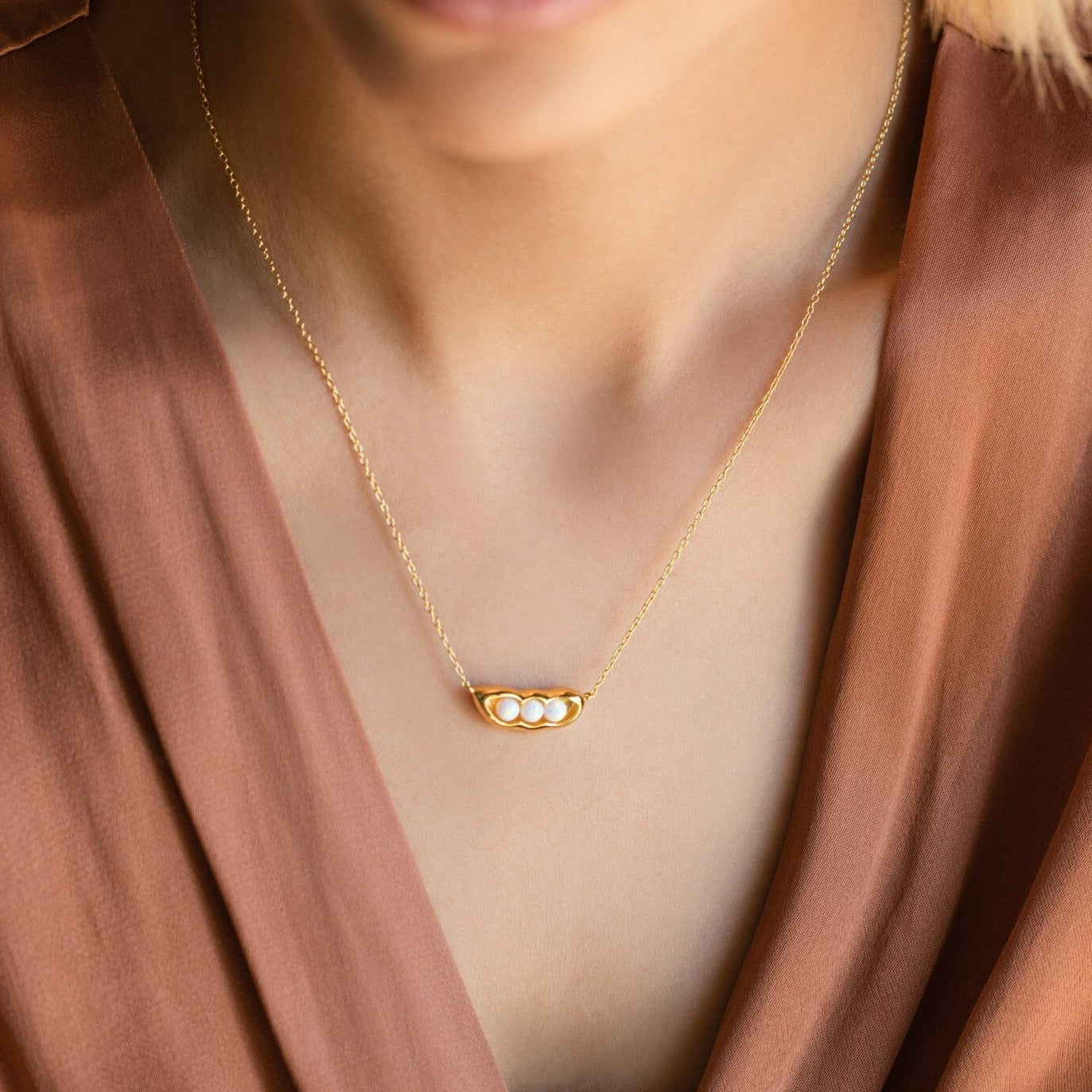 A woman wears the Peas in a Pod Necklace—a gold pendant with three pearls—paired with a brown V-neck top, highlighting meaningful jewelry.