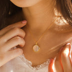 Woman wearing a Pearl Disc Locket Necklace with a round pendant, touching the chain and dressed in a white top—a meaningful gift and timeless accessory.