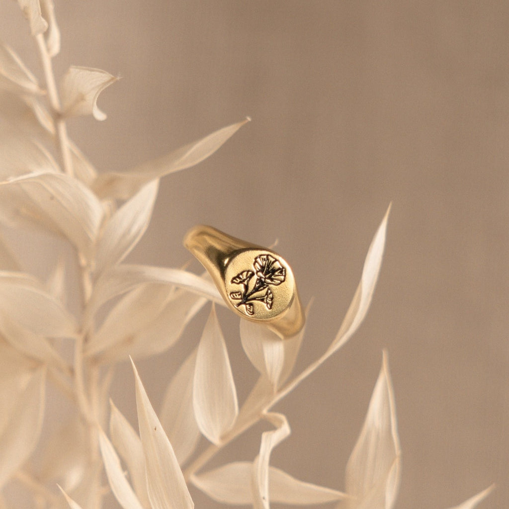 The Birth Flower Signet Ring features a delicate floral engraving and rests on pale dried leaves against a beige background—a charming piece of personalized jewelry.