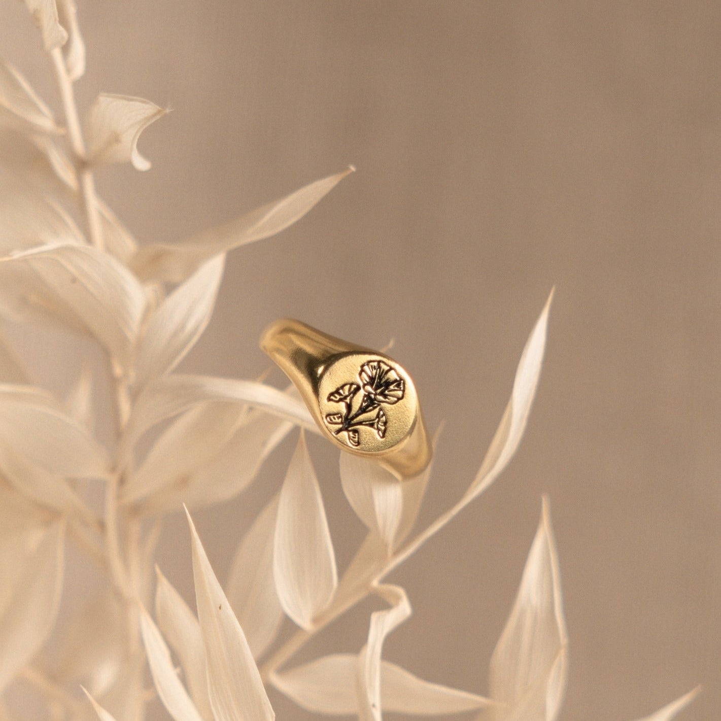 The Birth Flower Signet Ring features a delicate floral engraving and rests on pale dried leaves against a beige background—a charming piece of personalized jewelry.