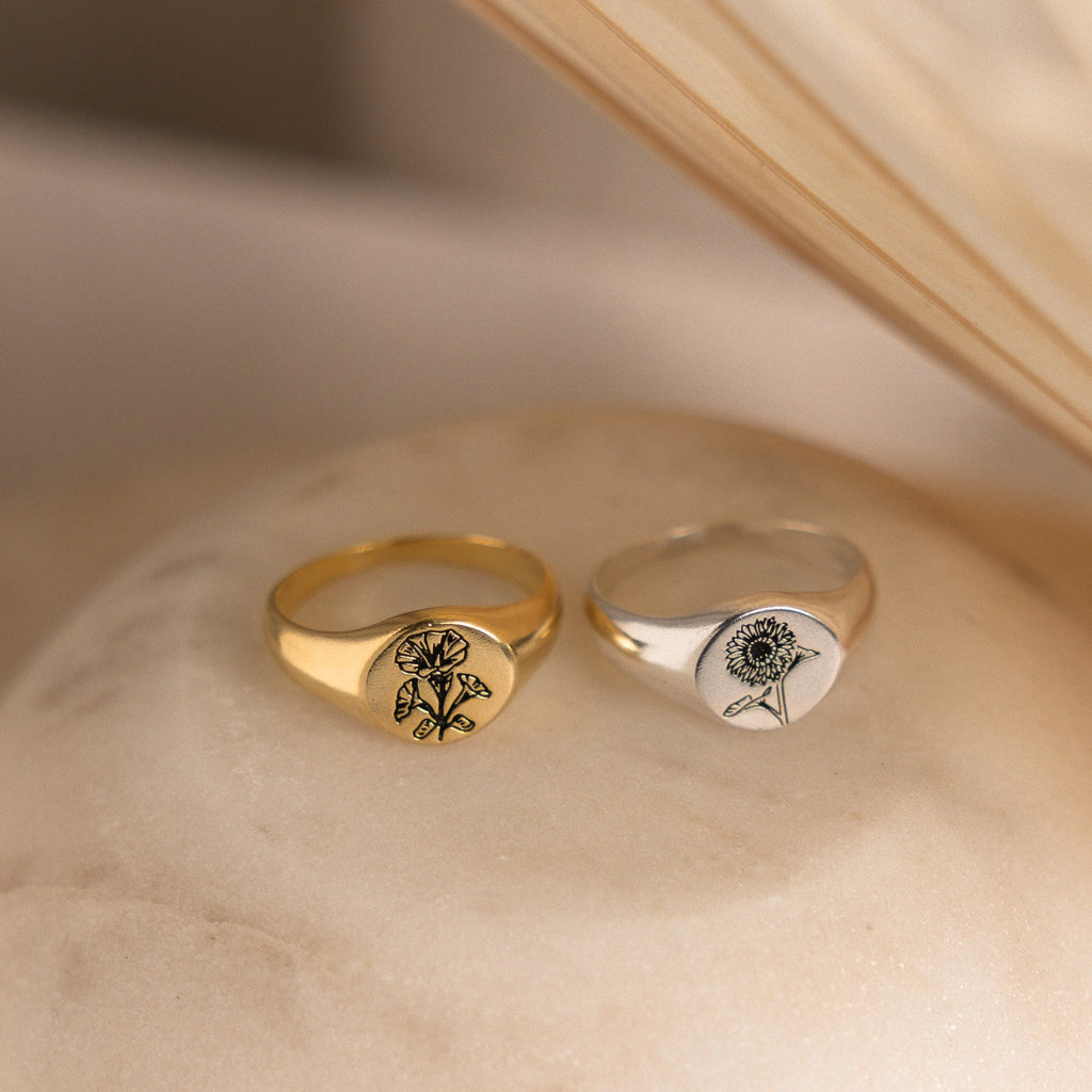 A Birth Flower Signet Ring rests on a soft beige surface, showcasing elegant, personalized jewelry.