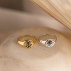 A Birth Flower Signet Ring rests on a soft beige surface, showcasing elegant, personalized jewelry.