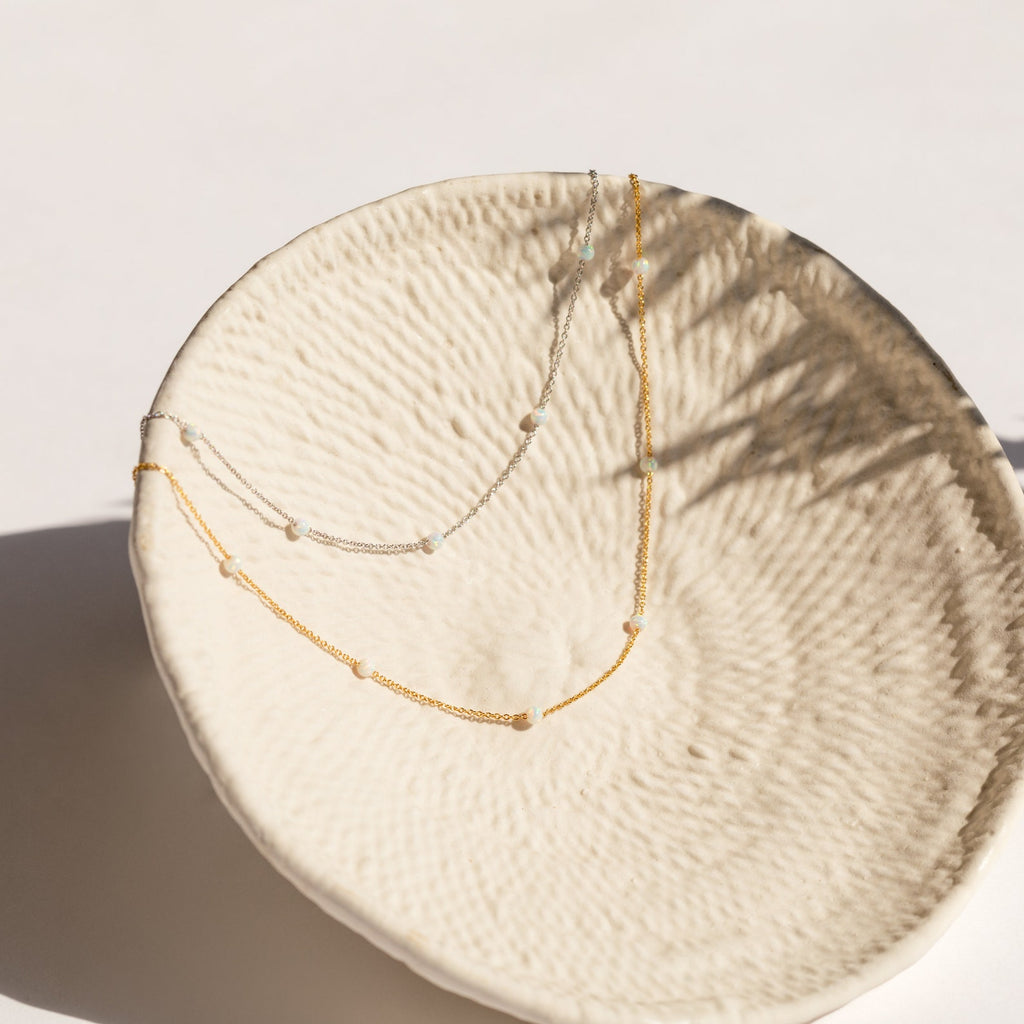 Gold and silver chain necklaces with evenly spaced small opal beads, displayed draped inside a textured cream ceramic bowl.