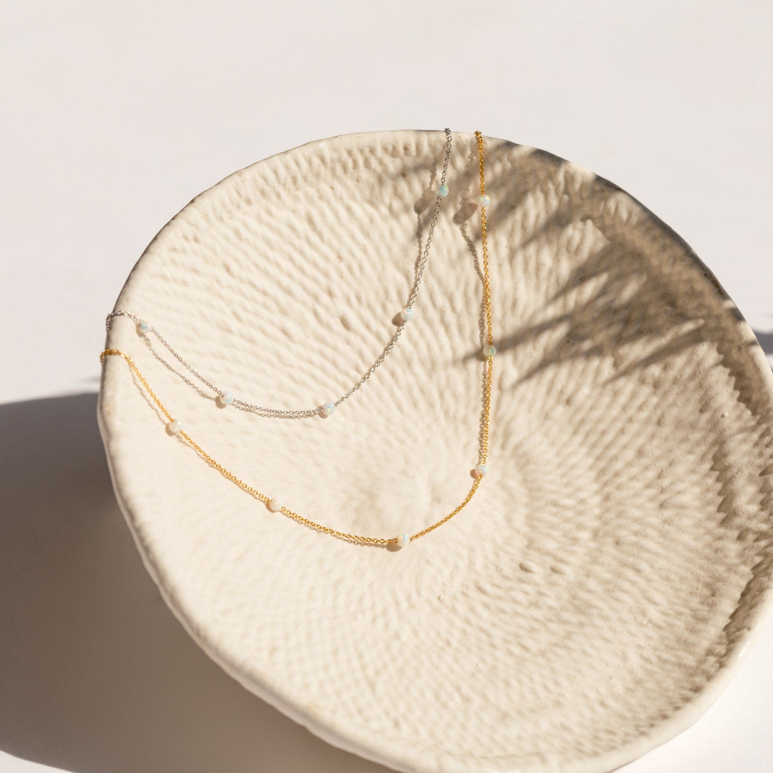 Gold and silver chain necklaces with evenly spaced small opal beads, displayed draped inside a textured cream ceramic bowl.
