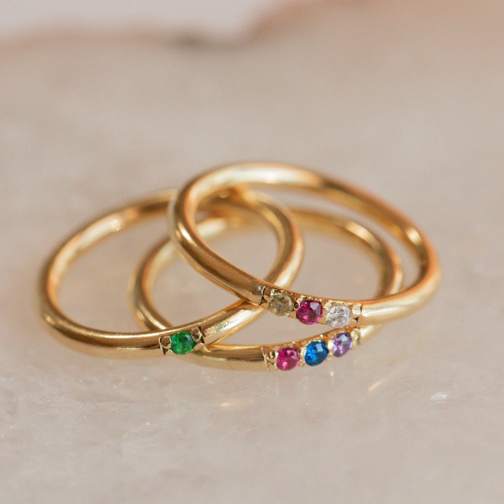 The Mini Birthstone Ring features a slim gold band set with vibrant gemstones, shown on a light marble surface—a delicate, elegant choice for everyday wear or as a personalized birthstone ring.