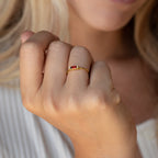 A woman's hand wearing the Empire Garnet Ring, showcasing a gold band with a small red baguette garnet, held near her face.