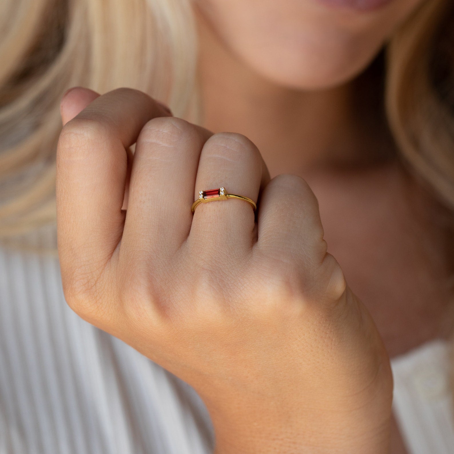A woman's hand wearing the Empire Garnet Ring, showcasing a gold band with a small red baguette garnet, held near her face.