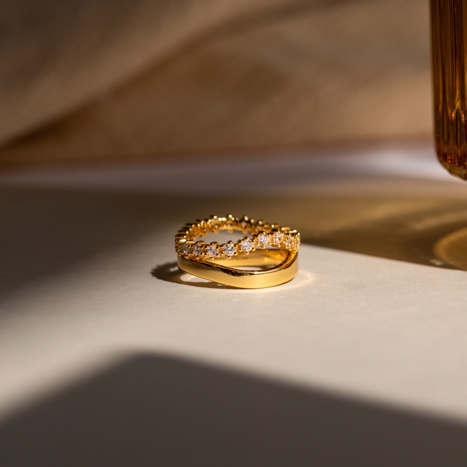 The Wave Diamond Duo Ring, featuring embedded gemstones, rests on a white surface illuminated by soft, warm light.