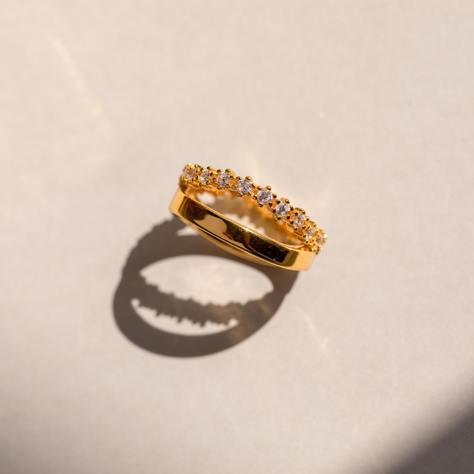 The Wave Diamond Duo Ring features a row of sparkling diamonds and casts an elegant shadow on a light surface.