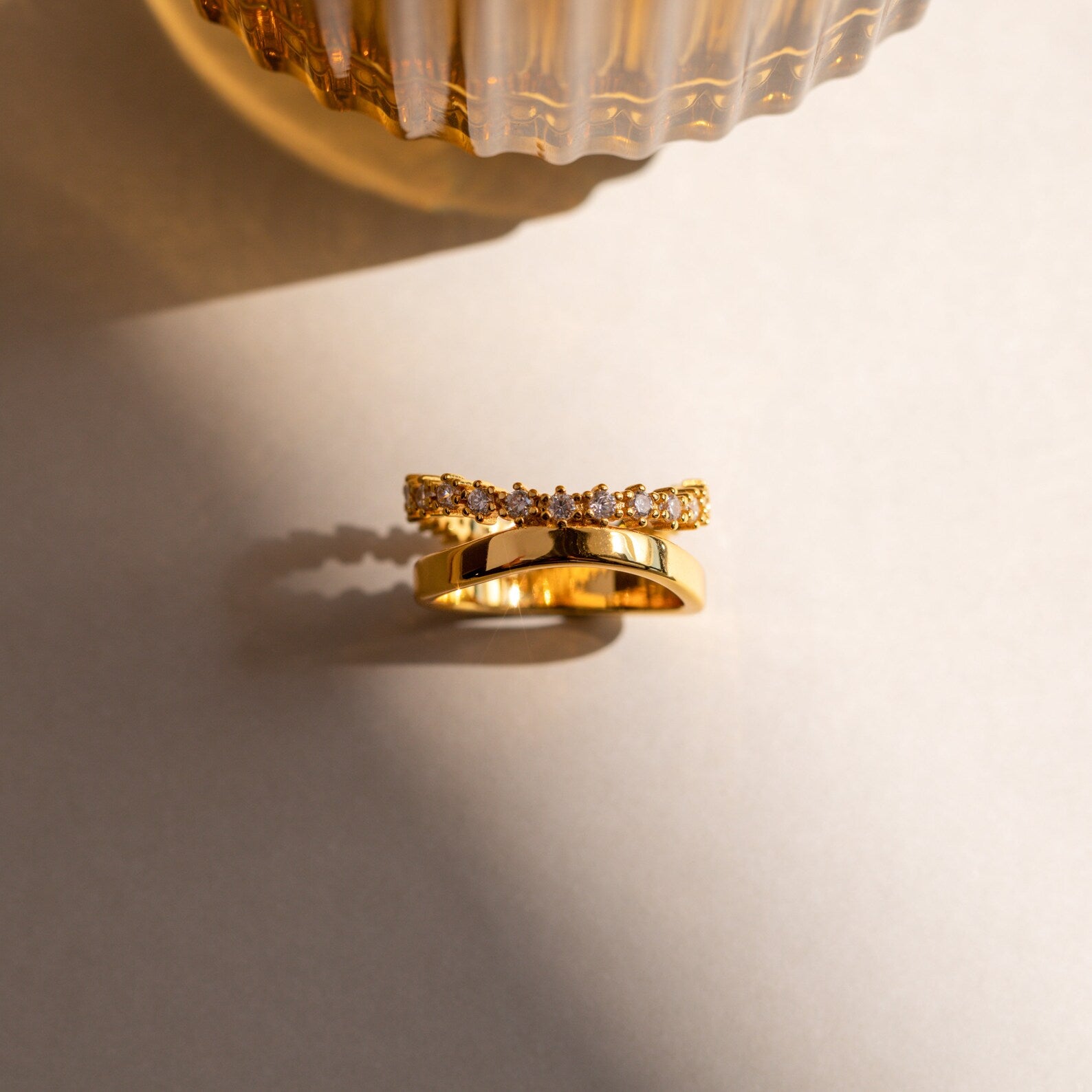 The Wave Diamond Duo Ring, a delicate gold piece adorned with small clear gemstones, rests on a neutral surface beneath glass decor and soft shadow.